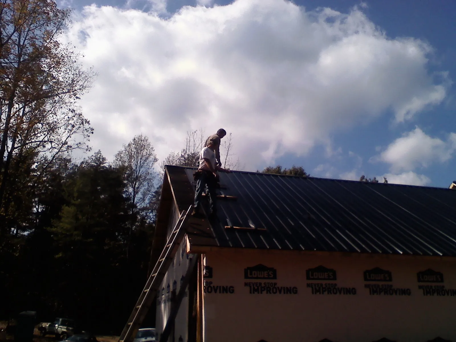 Roofer fastening dark metal panels high against a cloudy sky, from a Roof Inspection job in Saint Benedict